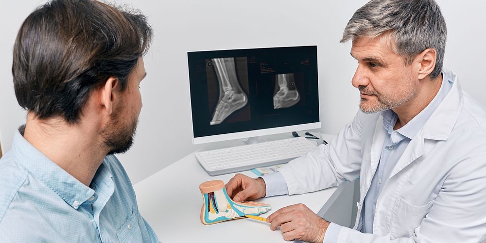 Ein Arzt erklärt einem Patienten die Ursachen eines Hallux Valgus anhand eines Fußmodells.