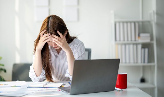 Eine Frau sitzt am Schreibtisch und fühlt wegen ihres Burnouts körperliche Symptome wie Kopfschmerzen.